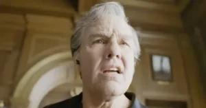 The image shows a person with white hair looking upwards in a grand, possibly historical, setting with arched architecture and a window in the background.