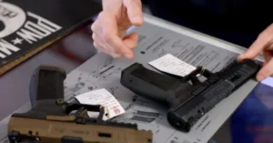 The image shows a close-up of a person's hands handling a disassembled handgun on a table with diagrams and instructions.