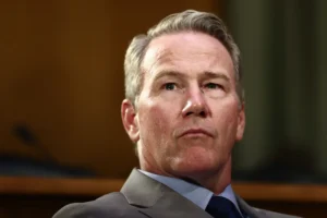 The image shows a man in a suit and tie, appearing to be in a formal setting, possibly during a professional or political event.