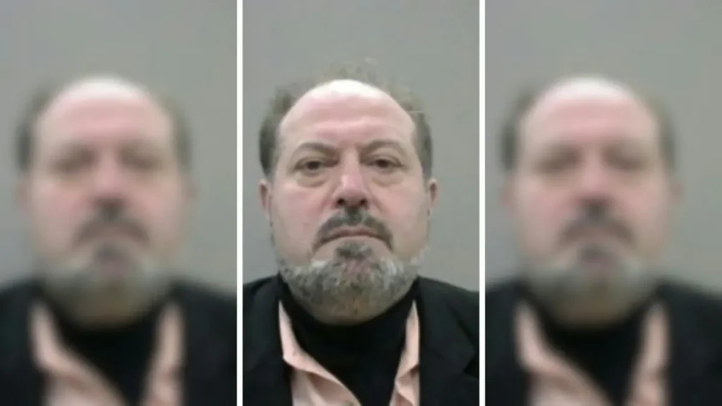 The image appears to be a mugshot of a man with a beard and mustache, wearing a black suit and a light-colored shirt.