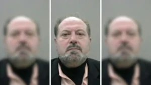 The image appears to be a mugshot of a man with a beard and mustache, wearing a black suit and a light-colored shirt.