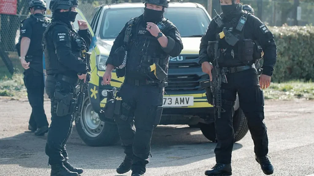 A group of armed police officers in tactical gear are seen standing near a police vehicle, ready for action.