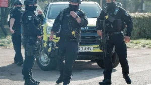 A group of armed police officers in tactical gear are seen standing near a police vehicle, ready for action.