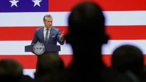 A man in a suit stands at a podium with the Department of War seal, delivering a speech in front of an American flag backdrop.
