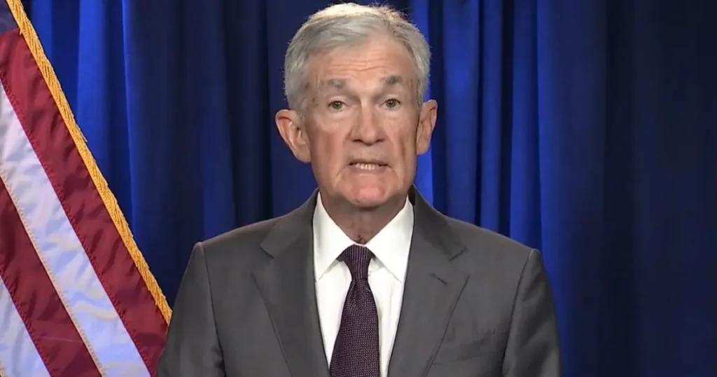 The image shows a man in a suit and tie speaking in front of a blue curtain and an American flag.