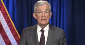 The image shows a man in a suit and tie speaking in front of a blue curtain and an American flag.