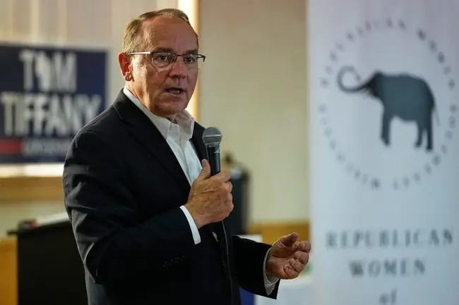 A man in a suit is speaking into a microphone at an event, with a sign for "Republicans Women" and an elephant logo in the background.