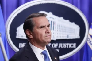 The image shows a man in a suit standing in front of the Department of Justice seal.