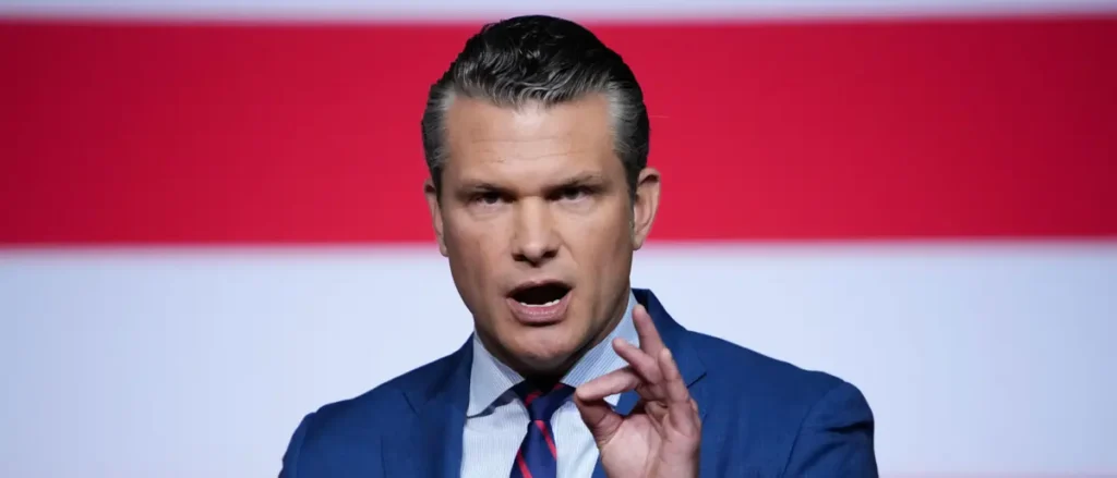 A man in a blue suit and tie is speaking in front of a red and white background.