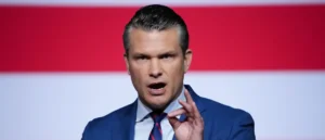 A man in a blue suit and tie is speaking in front of a red and white background.