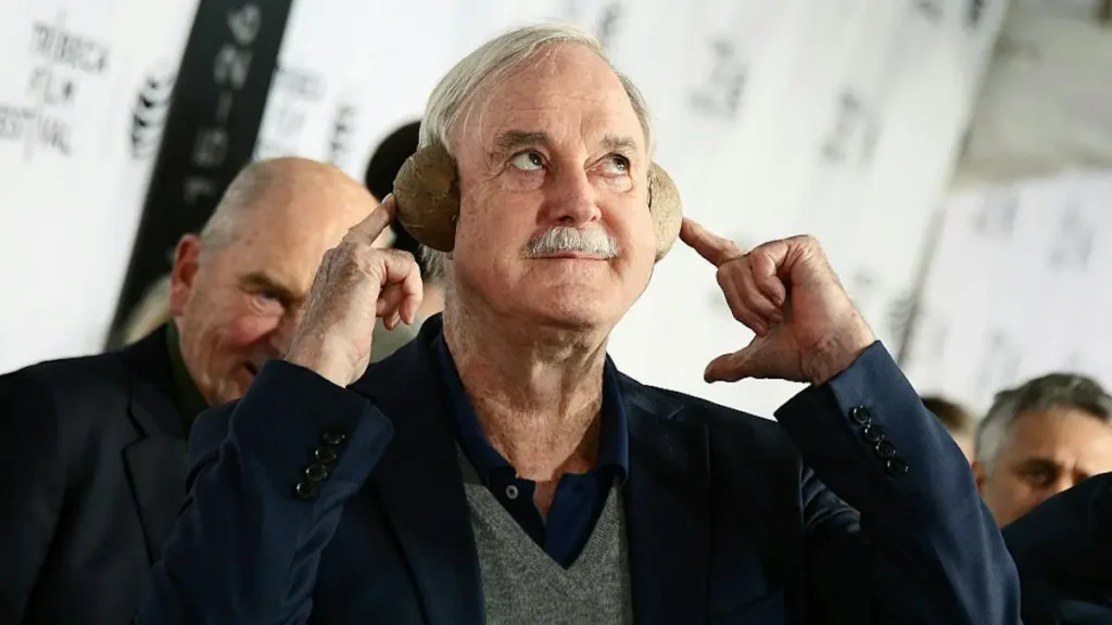 An older man adjusts his earmuffs at a public event, with another individual in the background.