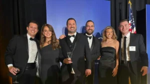 A group of six people, dressed in formal attire, pose for a photo holding an award at what appears to be a formal event.