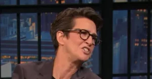 The image shows a person wearing glasses and a dark blazer, speaking on a television show set against a cityscape backdrop.