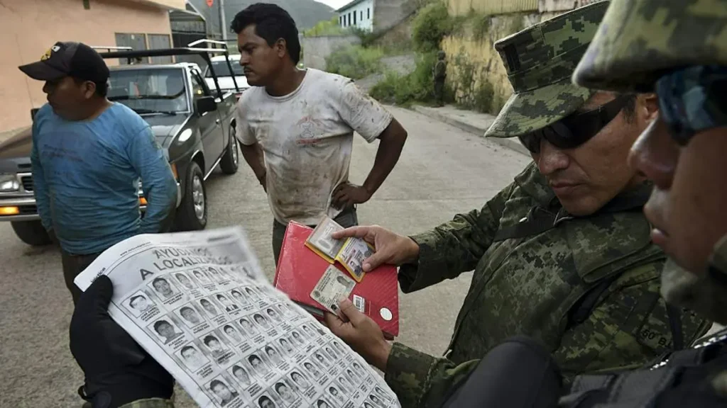A soldier examines identification documents while a man holds a poster with missing persons images in a street setting.