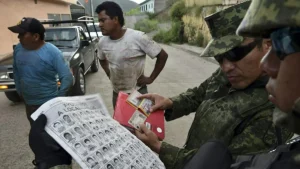 A soldier examines identification documents while a man holds a poster with missing persons images in a street setting.