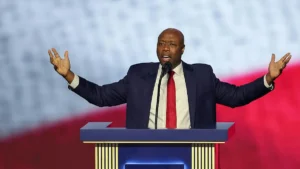 A man in a suit and tie stands at a podium with his arms outstretched, addressing an audience.