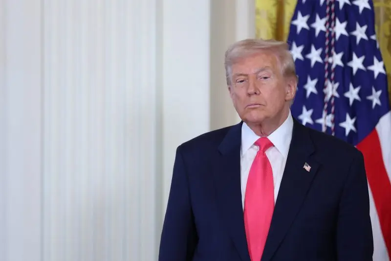 The image shows a man in a dark suit and red tie standing in front of an American flag.