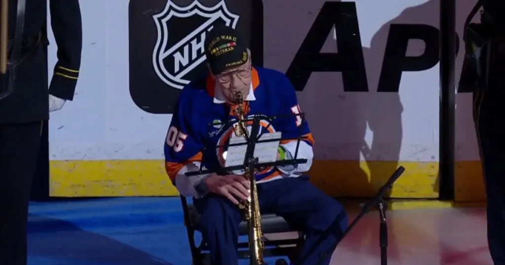 An elderly man in a hockey jersey plays a saxophone on stage during a ceremonial event.