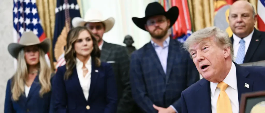 The image shows a group of individuals, including a man in a suit and tie, standing in front of American flags, with some wearing cowboy hats, in what appears to be a formal setting.
