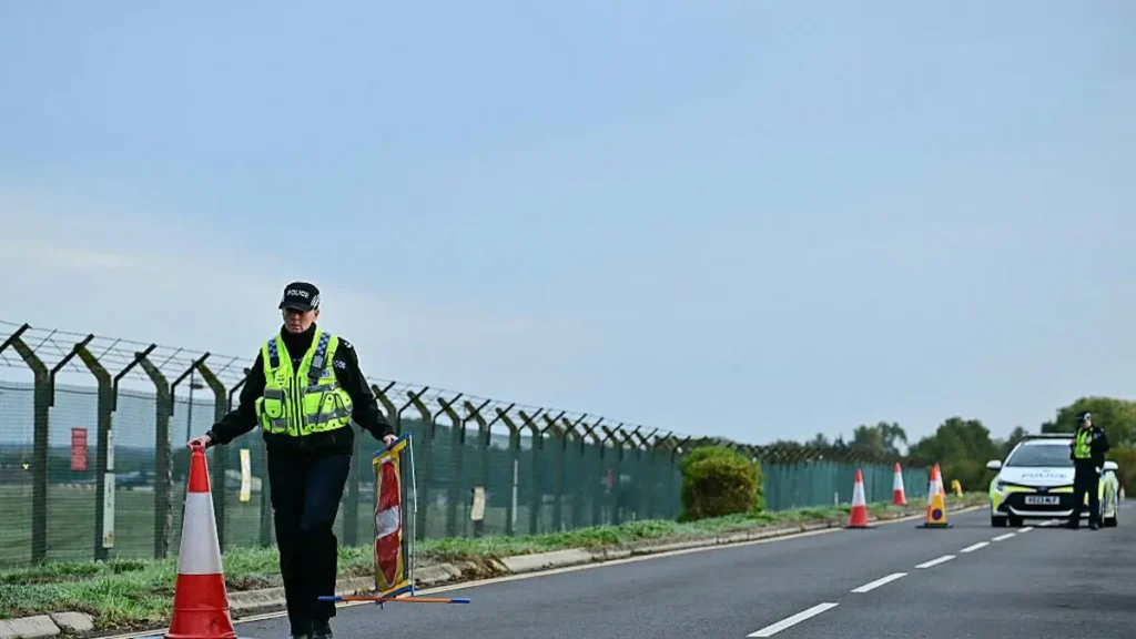 A police officer is directing traffic with cones and flags near a road with a fence and a police car in the background.