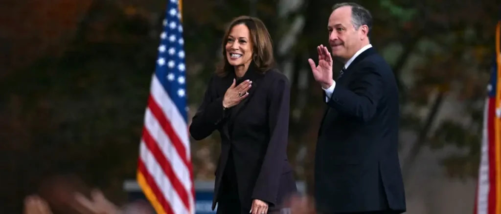 The image shows two individuals, one of whom is placing a hand over their heart, likely during a formal event or ceremony, with an American flag in the background.