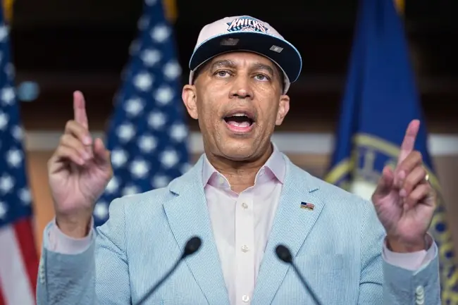 The image shows a person speaking at a podium with American flags in the background, wearing a light blue blazer and a cap with "Knicks" written on it.