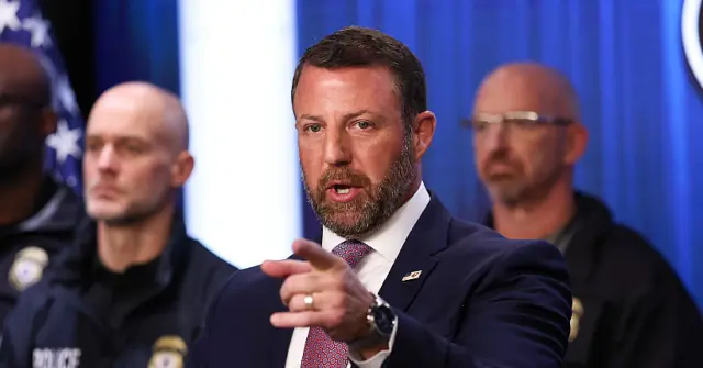 A man in a suit and tie is speaking at a press conference, pointing to the side, with two police officers in the background.
