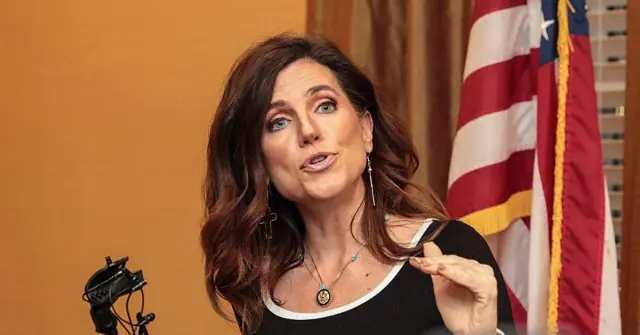 A woman is speaking in front of an American flag, gesturing with her hand.