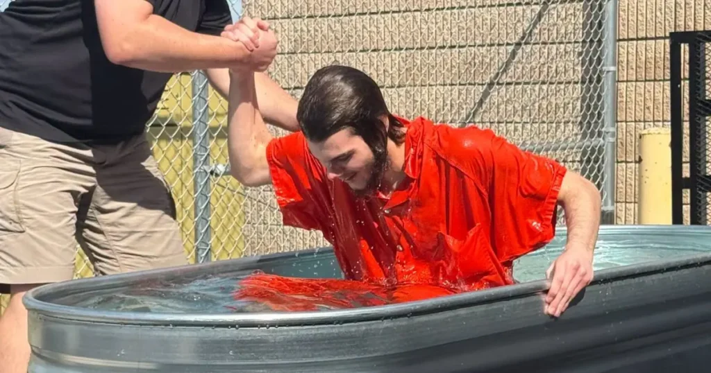 A person in a red shirt is being dunked into a large container of water, possibly as part of a baptism or similar ritual.