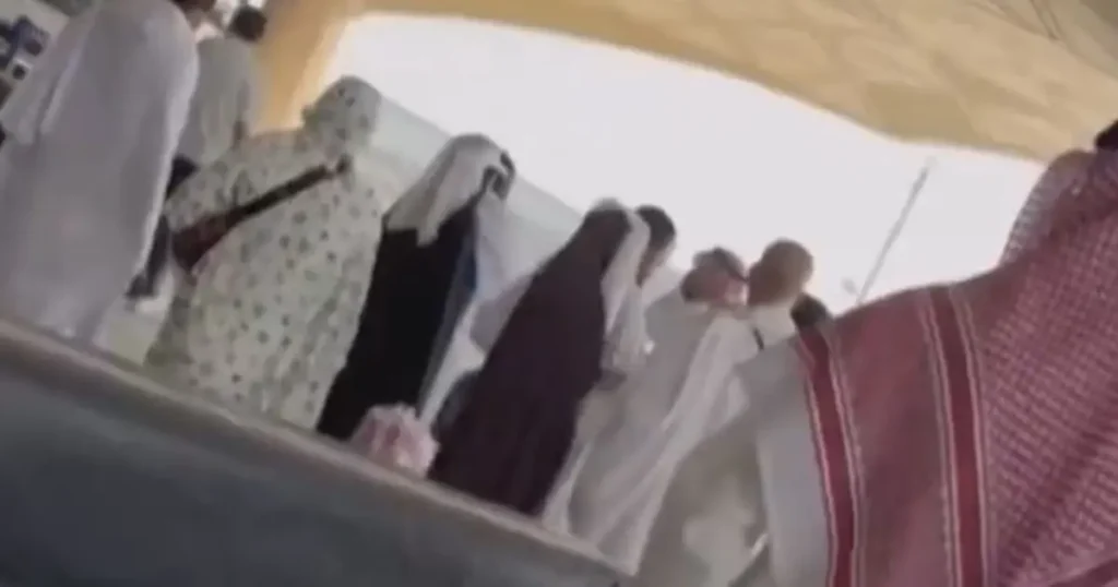 The image shows a group of people, some wearing traditional attire, gathered under a tent-like structure, possibly at a cultural or religious event.