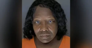 The image is a mugshot of a person wearing an orange shirt, with a plain background.
