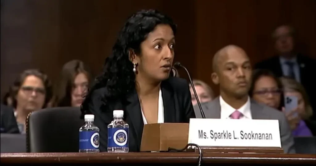 A woman is speaking at a formal hearing, with a nameplate in front of her that reads "Ms. Sparkle L. Sooknanan."