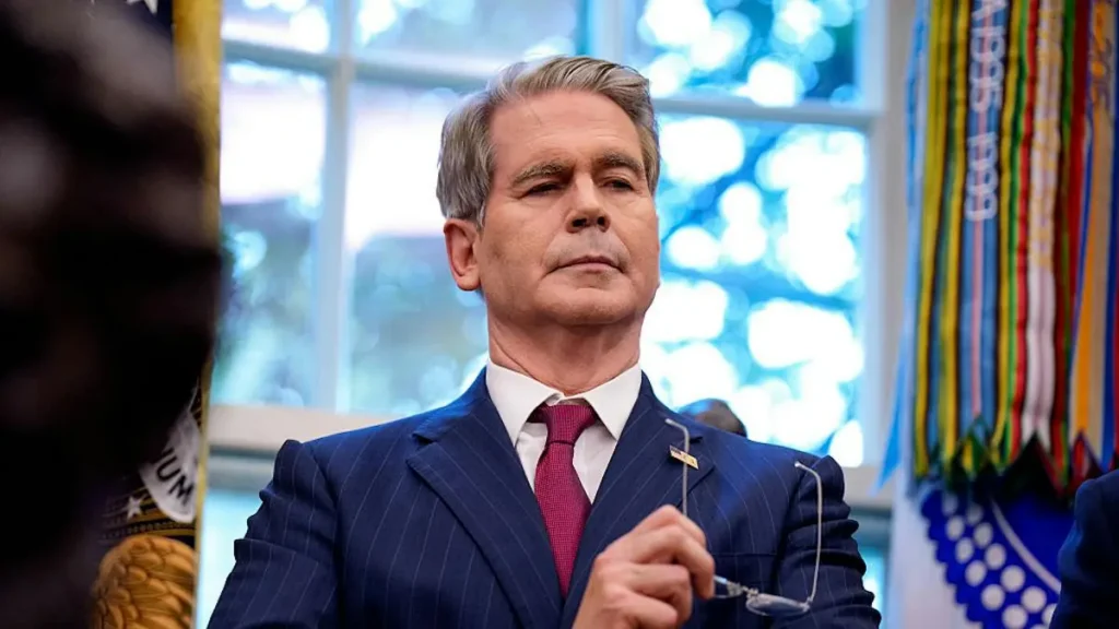 The image shows a man in a formal suit and tie, holding a pair of glasses, in a setting with a window and decorative elements in the background.