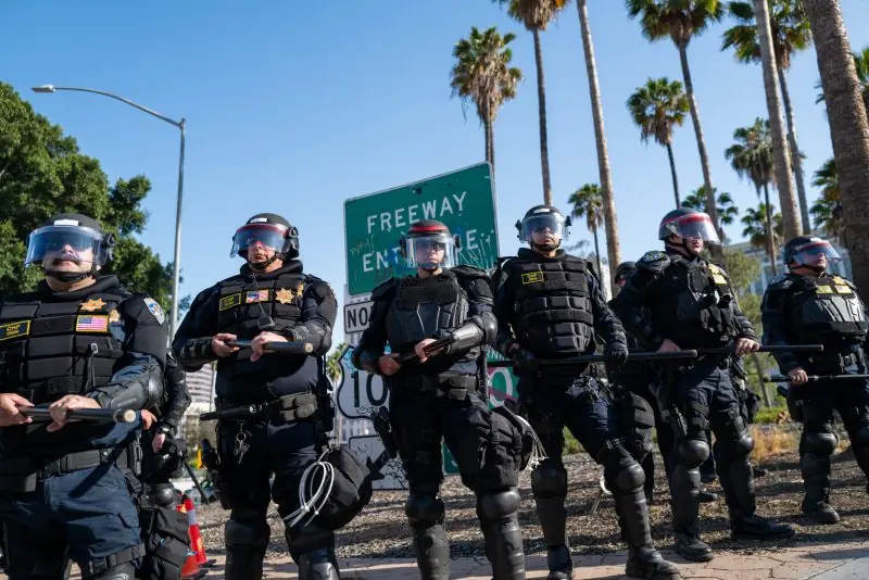A line of police officers in riot gear stands in formation near a freeway entrance sign.