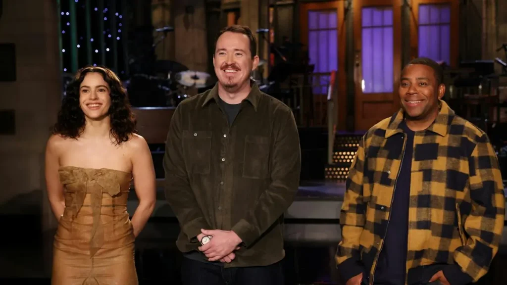 Three individuals are standing together in a studio setting, smiling and posing for the camera.