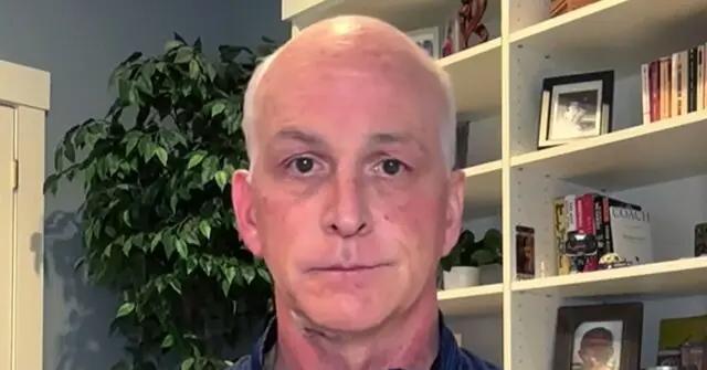 The image shows a man in a home office setting, with a bookshelf and a potted plant in the background.