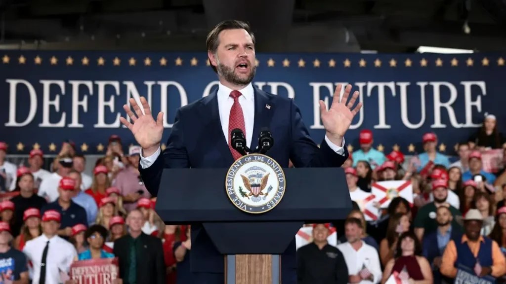 A politician stands at a podium with a "Defend Our Future" backdrop, addressing a crowd of supporters.
