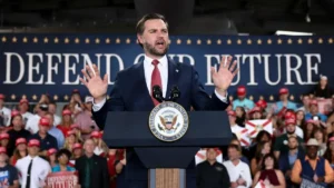 A politician stands at a podium with a "Defend Our Future" backdrop, addressing a crowd of supporters.