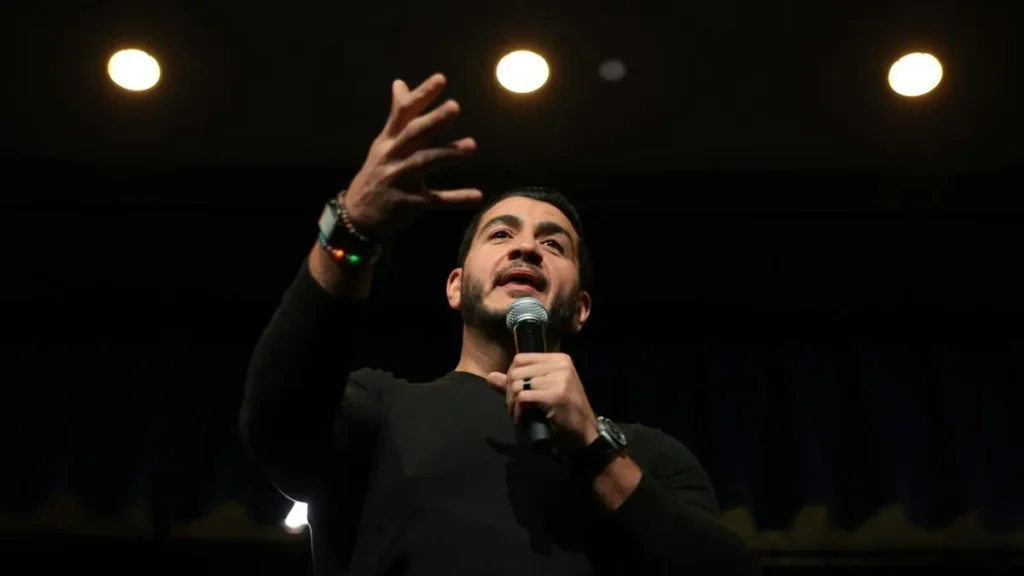 A man is speaking into a microphone while gesturing with his hand, likely during a public speaking event or performance.