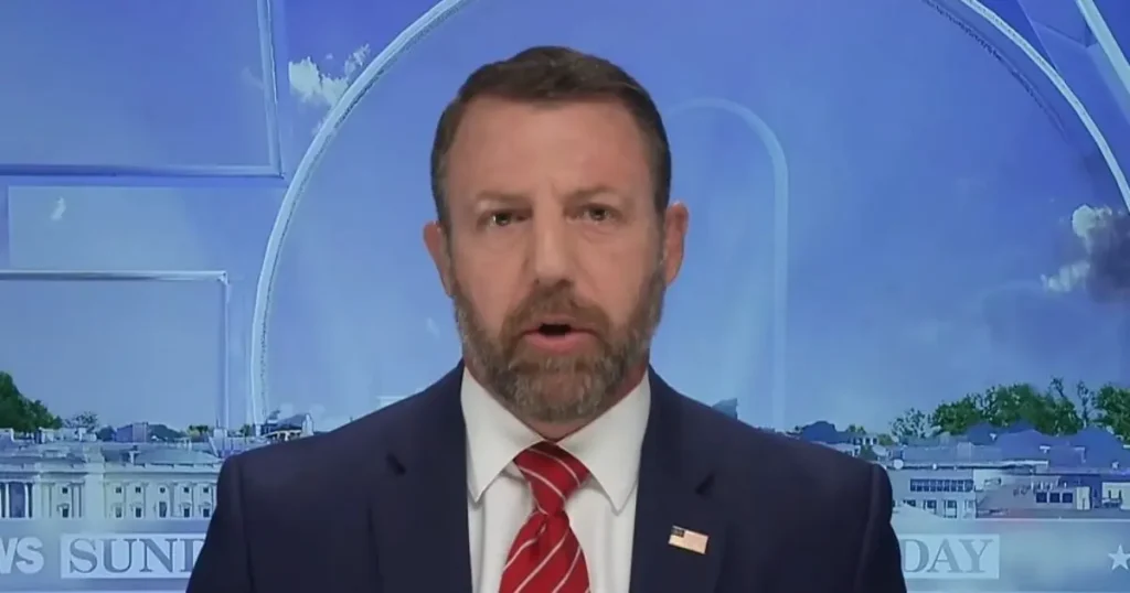 The image shows a man in a suit and tie, speaking on a news program with a backdrop featuring the Washington Monument and the White House.