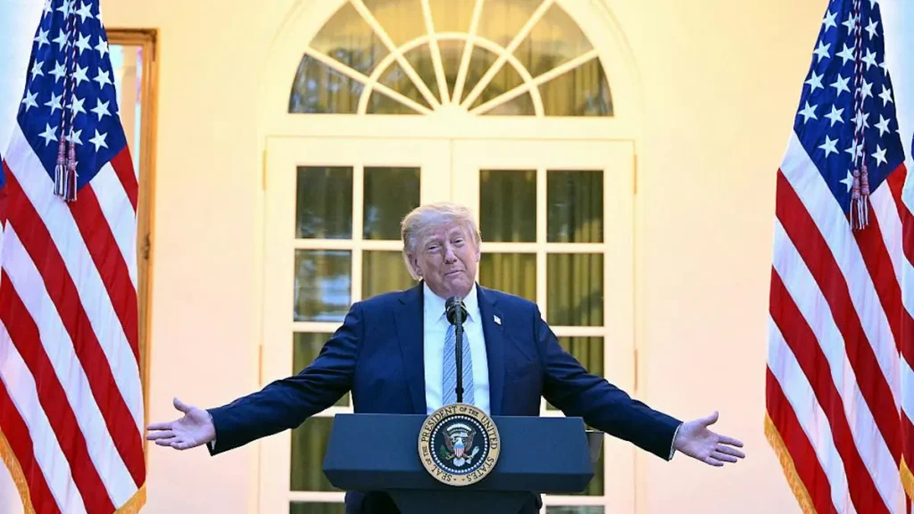 The image shows a person standing at a podium with the seal of the President of the United States, flanked by two American flags, in front of a door with a fanlight window.