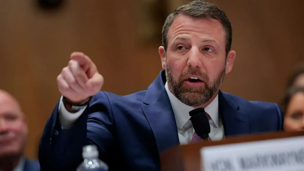 A man in a suit is speaking at a formal event, gesturing with his right hand.