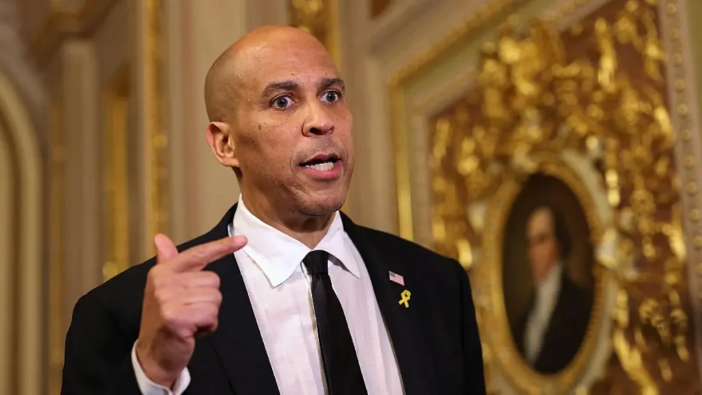 The image shows a man in a suit and tie, gesturing with his hand, in a formal setting with ornate gold decorations and a portrait in the background.