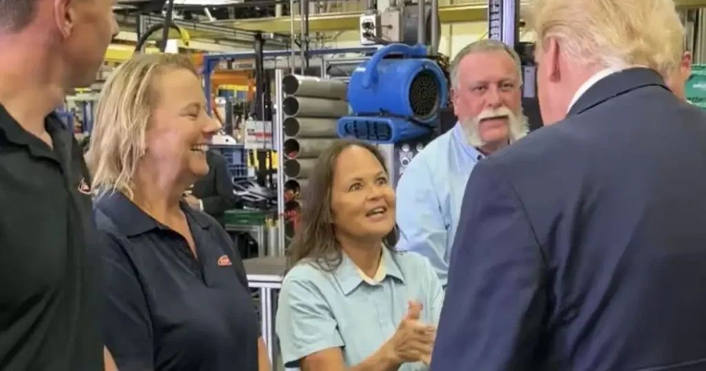 The image shows a group of people in a factory setting, engaging in conversation with a man in a suit, who appears to be a visitor or official.