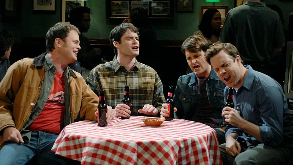 Four men are gathered around a table with beer bottles and a bowl, appearing to be engaged in a lively conversation in a bar setting.