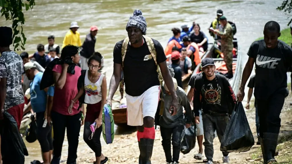 A group of people, some carrying bags, are walking along a riverbank, possibly after being rescued or relocating.