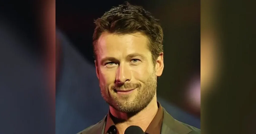 The image shows a man with a beard and short hair, smiling while speaking into a microphone.