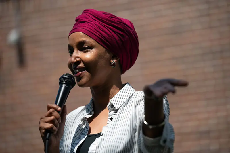A woman wearing a red turban speaks into a microphone while gesturing with her hand.