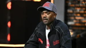 The image shows a person wearing a black jacket with red text and a matching cap, sitting in a studio setting with blurred lights in the background.