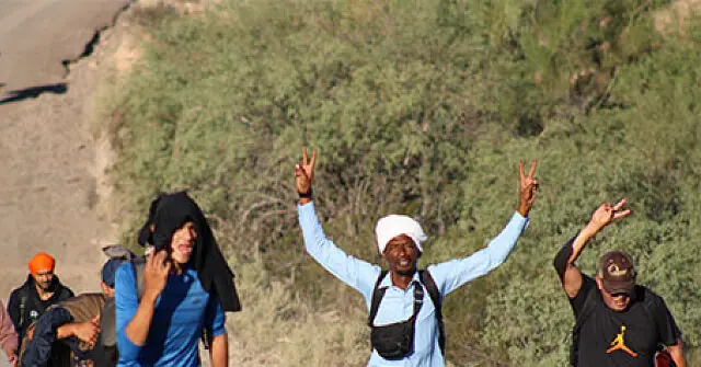 A group of people, including a man in a white turban, raise their hands in a peace sign while walking along a road.